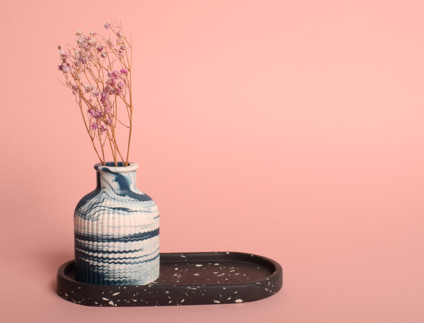 An oval-shaped black trinket tray with white terrazzo chips. The trinket tray is holding a blue and white marble effect vase that has dried flowers in it.